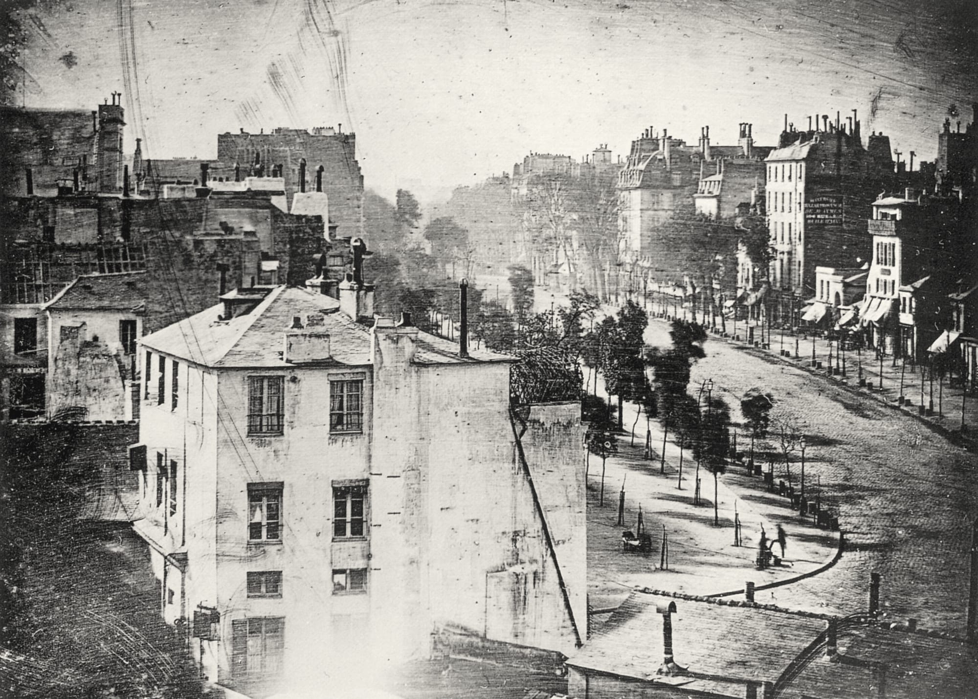 Historic black-and-white image of a 19th-century city Parisian street, with old buildings and trees lining the road. A lone figure is visible on the pavement.